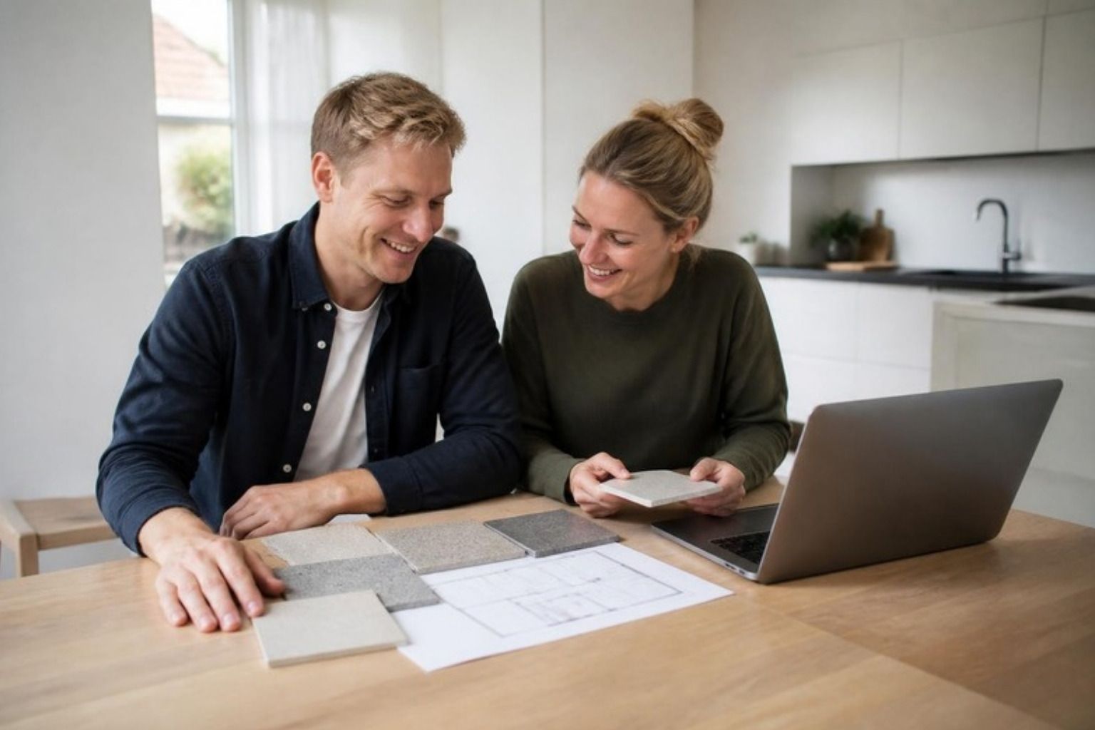 Smilende par ved et køkkenbord med fliseprøver, en plantegning og en bærbar computer